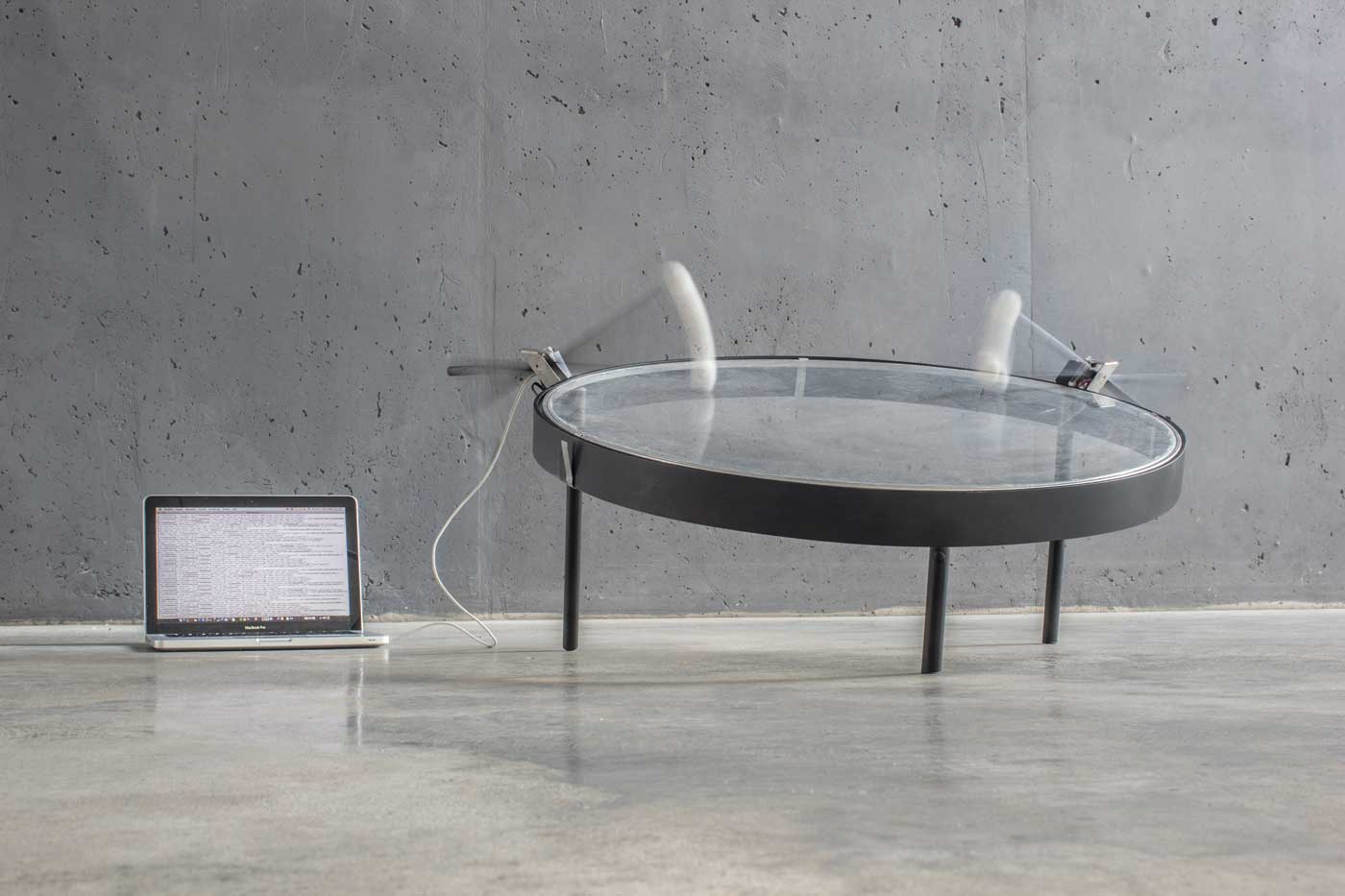 An exhibition photo, a laptop is connected to a large transparent drum frame being struck by mallets.