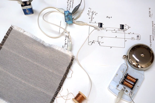 On top of a patent circuit illustration is a woven fabric, and a circuit consisting of a 9V battery connected by alligator clips to an electrical box and a bell.